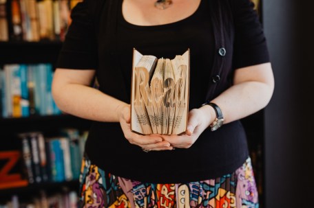 Torso of a woman wearing a black cardigan and brightly patterned skirt. She is holding a book which has been folded into the word READ. She is standing in front of a bookcase.
