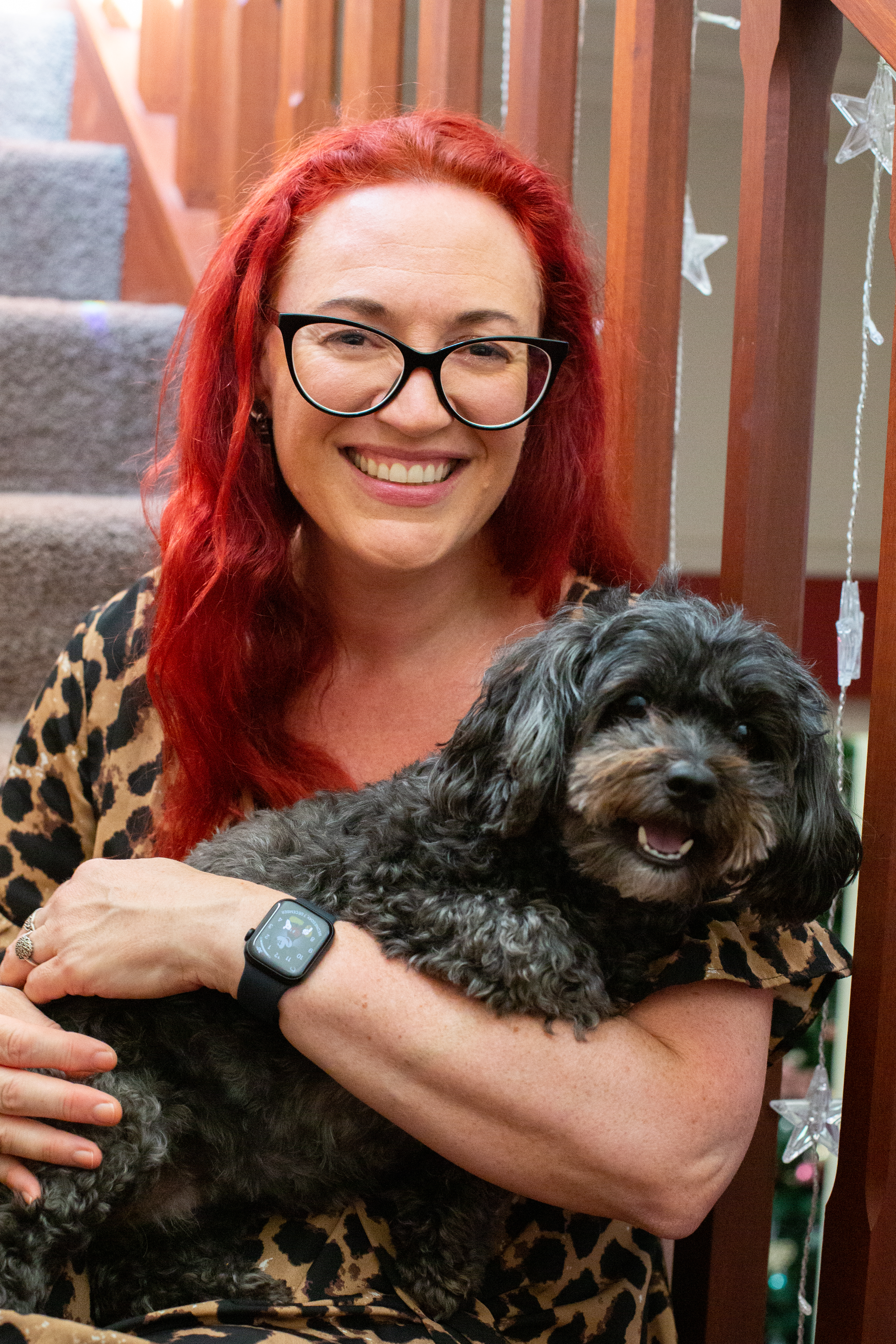 Smiling red headed woman with glasses sitting with small smiling black dog. Image credit JJ Gately Photography